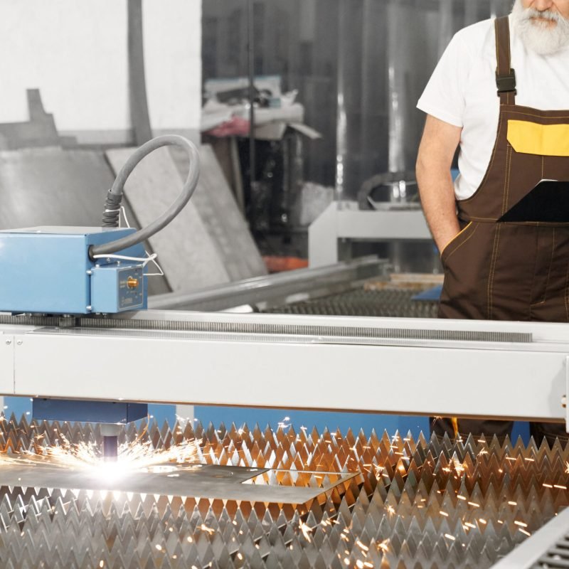 CNC laser plasma cutting metal, sparks. Engineer standing behind and observing process of plasma torch cutting steel sheet. Worker wearing in coveralls, white t shirt, holding folder.